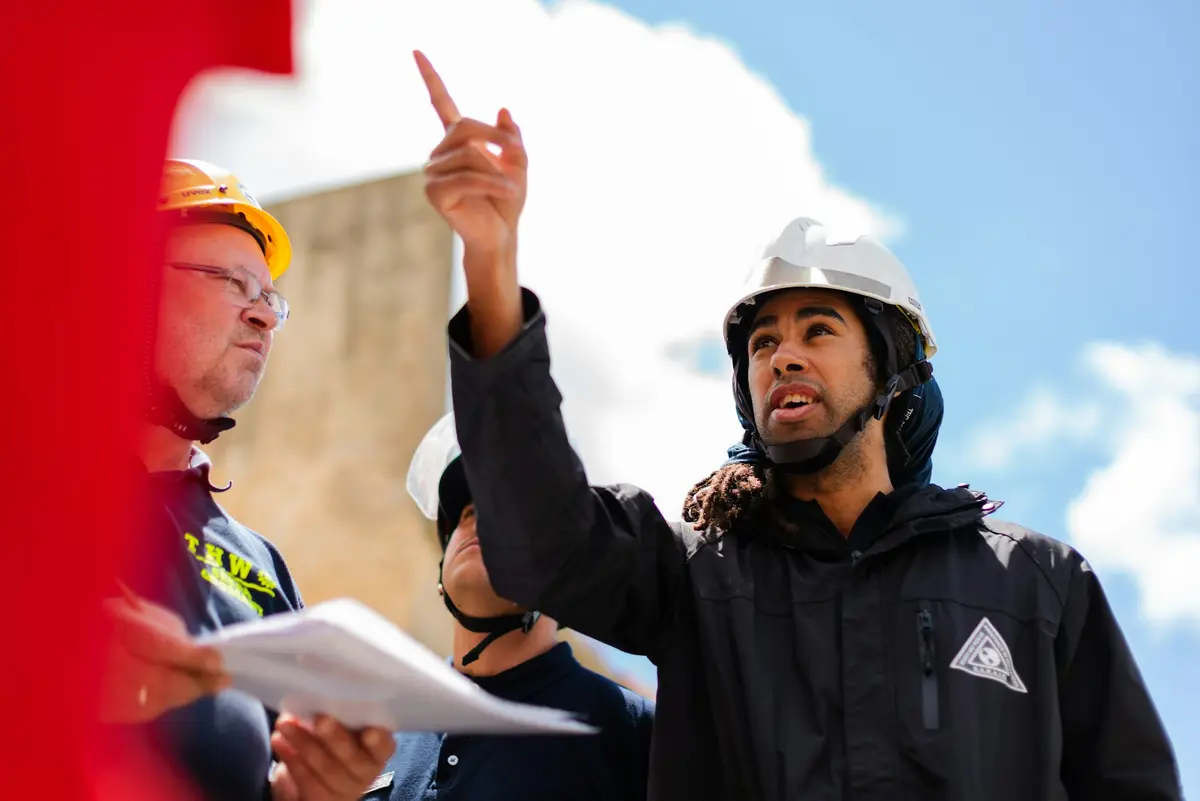 Engenheiros de capacete em uma obra, um deles apontando para cima enquanto o outro segura o projeto