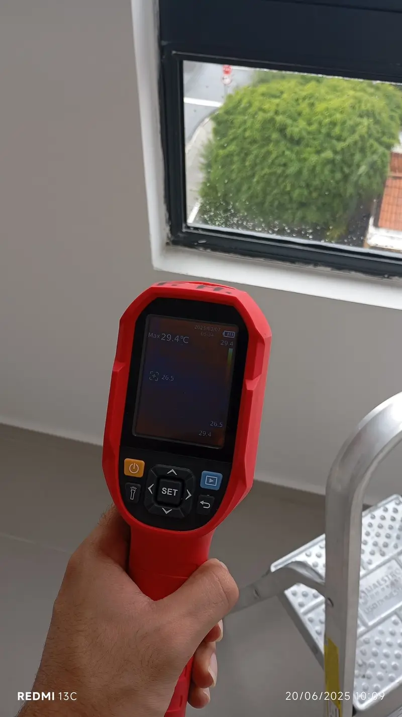 Mão segurando uma câmera termográfica apontada para a parede de um apartamento, com a imagem térmica na tela